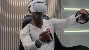Fütürizm oyun interneti video oyunu sanal gerçekliği VR gözlüklü Afrikalı Amerikalı erkek adam beyaz fütüristik odada 3 boyutlu dünya oynuyor kapalı mekanlarda modern internette eğleniyor metaverse oynamaktan zevk alıyor.