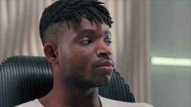 Afrikalı sakince gülümseyen, kameraya bakan siyah sandalyede oturan, kaygısız Amerikan erkek portresi modern odada fütürizm stili fütürizm yüz ifadesi siber konseptinde.