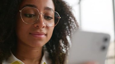 Etnik Afro-Amerikan kadın neşeli kız cep telefonu cihaz bağımlısı kız öğrenci olumlu gülümseyen mesajlar okuyor akıllı telefon gülümseme kullanıcısı elinde cep telefonu web sitesi var