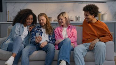 Evdeki beyaz adam, komik video cep telefonu arkadaşlarını kanepeye oturtup, çeşitli etnik gruplara mensup erkeklere gülüyor. Afrikalı erkekler, esprili sohbetler, eğlenceli sohbetler, kapalı mekan arkadaşlığı.