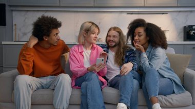 Çok ırklı grup dört arkadaş koltukta oturmuş birlikte gülüyor Kafkasyalı erkekler Afro-Amerikalı kadınlar haber cep telefonu uygulamasını tartışıyorlar kapalı alandaki mutlulukla buluşuyorlar