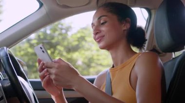 Gülümseyen Afrikalı Amerikalı iş kadını cep telefonunu araba şehrinin içinde kullanıyor neşeli iş kadını, etnik kız sürücü elinde cep telefonu, internette sosyal medya otomobili otoparkında geziniyor.