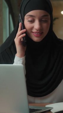 Neşeli gülümseyen cep telefonlu Müslüman kadın tesettürlü iş kadını Arap kadın çoklu iş kadını yönetici ofis ajansında çalışan dizüstü bilgisayar görünümlü iş kadını proje düzenini tartışıyor