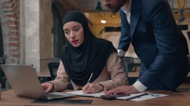 Kafası karışmış bir stajyer Müslüman iş kadını ve dizüstü bilgisayarlı bir iş adamı lideri iyi işi kontrol ediyor iş adamı desteğini kontrol ediyor mutlu bir kadına notlar yazıp işbirliği yapan bir takım çalışması yapıyor.