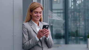 Gülümseyen Kafkasyalı orta yaşlı iş kadını cep telefonu tutuyor şirket binasının duvarına yaslanıyor gülümsüyor mutlu 40 'ların iş kadını şehirde e-posta sohbet akıllı telefonu kontrol ediyor.