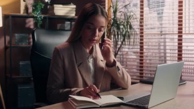 Kafkasyalı iş kadını cep telefonuyla konuşuyor. Laptop yazıyor. Ofis masasında birden fazla iş yapıyor.