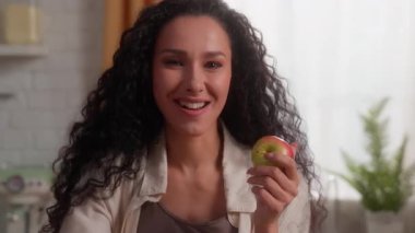 Mutlu, gülümseyen Latin bir kadın mutfakta taze elma yiyor. Neşeli bir kadın. Meyve yiyor, sağlıklı besleniyor.