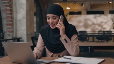 Mutlu Müslüman iş kadını telefon görüşmesi ile iletişim kuruyor. Laptop yazıları kullanarak tesettürlü Arap kadın akıllı telefon danışmanı. Şirket ofisindeki kaygısız, neşeli bayan kız.