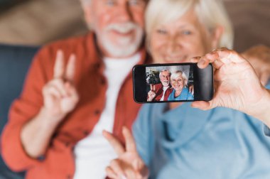Modern yaşlı insanlar. Kafkasyalı yaşlı çift koltukta oturup gülümserken cep telefonuyla selfie çekiyor.