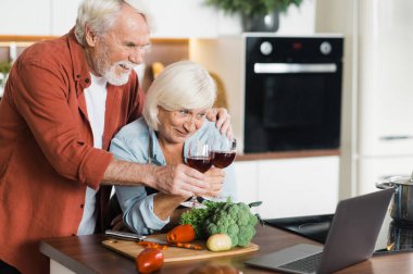 Mutlu, gri saçlı eşler mutfakta bir bardak şarapla ayakta duruyor ve evdeki mutfakta dizüstü bilgisayar kullanarak aile veya arkadaşlarıyla görüntülü görüşme yapıyorlar.