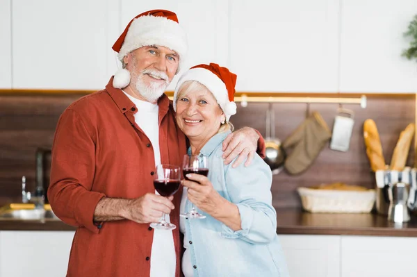 Portrait of elderly gray-haired couple in love standing in the kitchen in Christmas hats and with a glass of wine in their hands and looking at the camera with a smile