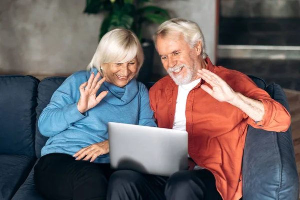 Elderly modern spouses communicate by video call using a laptop with family or friends while sitting at home on the couch and waving their hands to the camera