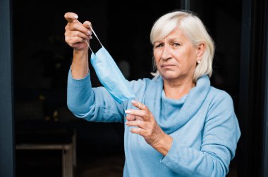 Olgun, gri saçlı, beyaz bir kadın elinde tıbbi bir maskeyle kameraya bakıyor.