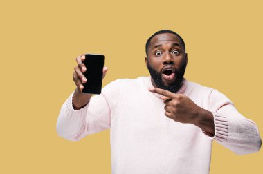 Sıradan giyinmiş Afrikalı Amerikalı adam izole edilmiş turuncu arka planda duruyor ve kameraya bakarken şaşkınlıkla telefonu işaret ediyor.