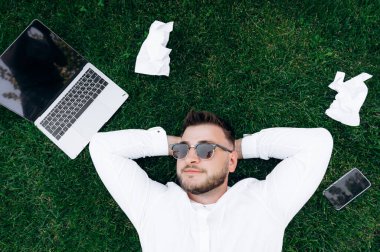 Çimlerin üzerinde çalışırken ara verin. Beyaz gömlek ve güneş gözlüğü takmış, çimenlerin üzerinde uzanmış, kendine güveni tam beyaz bir iş adamı. Yakınlarda bir dizüstü bilgisayar, telefon ve buruşuk çalışma kağıtları var.