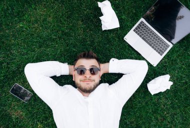 Çimlerin üzerinde dinleniyor. Beyaz gömlekli ve güneş gözlüklü mutlu beyaz adam çimenlerin üzerinde yatarken dinleniyor. Yakınlarda bir dizüstü bilgisayar, telefon ve buruşuk çalışma kağıtları var. Üst görünüm