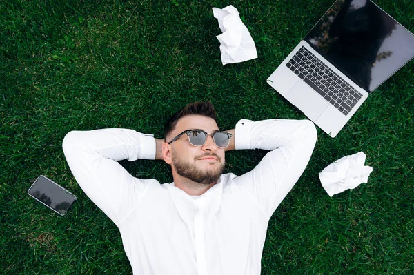 Handsome guy on the grass. Young attractive guy in a white shirt and sunglasses resting from work while lying on the lawn. Nearby lies a laptop, phone and crumpled work papers