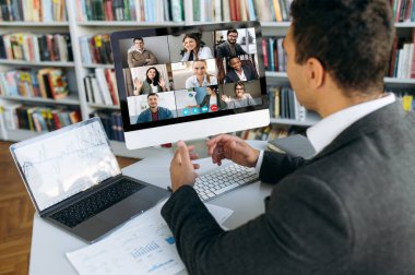 Online video meeting. Successful young adult businessman communicate with  business colleagues by video call using computer discussing about financial graphs and strategy clipart