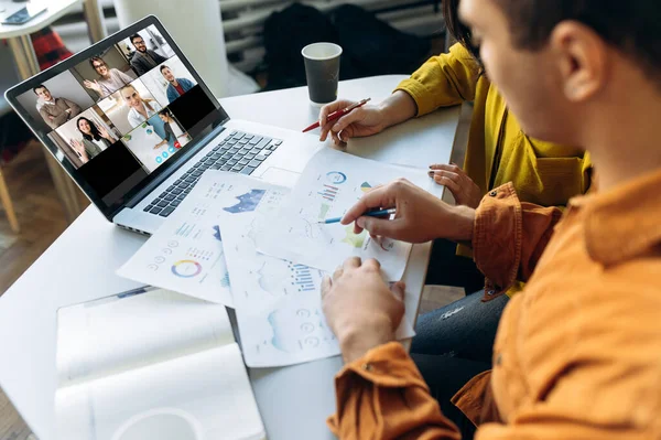 Online video chat. Laptop monitor view with multiracial business colleagues discussing about financial graphs, man and woman sitting on their workplace during video conference