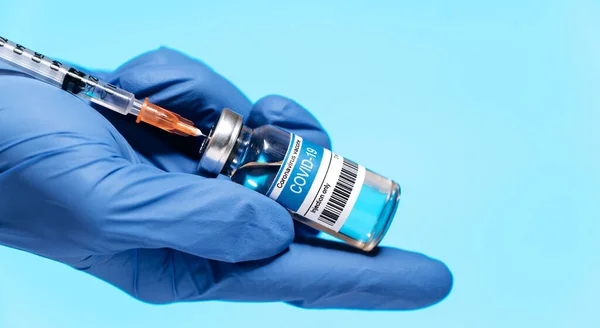 Close-up panoramic photo of a doctor\'s hand in medical glove holding syringe and ampoule with COVID-19 corona virus vaccine  injection. Coronavirus prevention, healthcare concept. Blue background