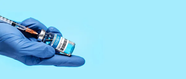 Panoramic photo of a doctor\'s hand wears medical glove holding syringe and ampoule with COVID-19 corona virus vaccine  injection. Coronavirus prevention, health and medical concept. Blue background