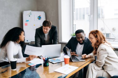 İş brifingi toplantısı. Finansal grafiklere ve diyagramlara bakan başarılı bir grup insan birlikte beyin fırtınası yapıyorlar. Çok ırklı erkek ve kadın çalışanlar yeni projeler üzerinde çalışırken fikirleri tartışıyor.