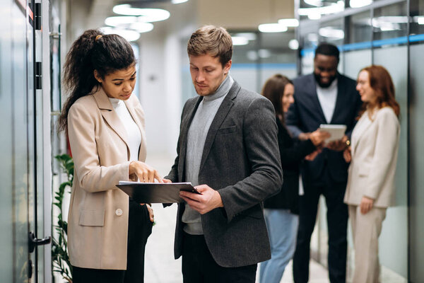 Multiethnic business colleagues stand in office. Beautiful african american female employee consults caucasian male colleague on working process. Focused young adult woman and man discuss the project