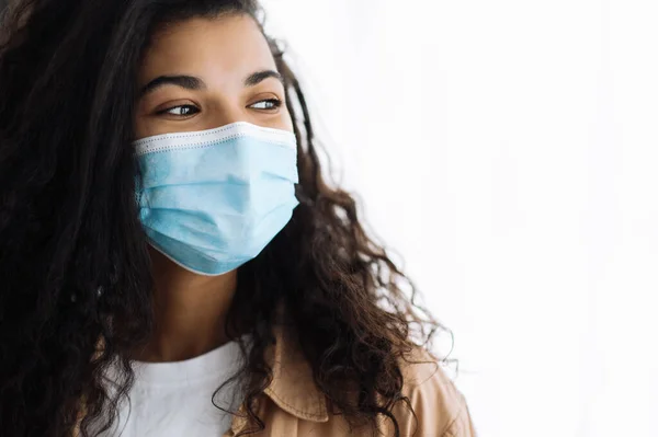 Medidas de prevenção. Africano americano bonita mulher morena em casual desgaste em pé dentro de casa com máscara médica protetora em seu rosto e olha para o lado — Fotografia de Stock