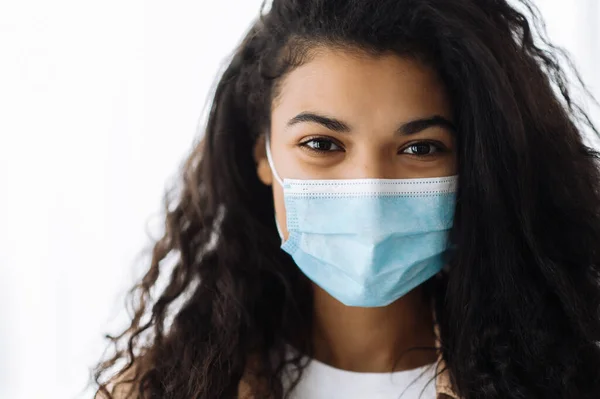 Close-up retrato de Africano americano atraente mulher morena em roupas casuais em pé dentro de casa usando uma máscara médica protetora em seu rosto e olhando diretamente para a câmera — Fotografia de Stock