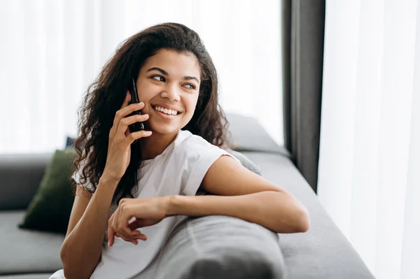 Seorang gadis cantik yang bahagia duduk di sofa di rumah. Wanita muda yang menarik berbicara melalui telepon dengan teman, keluarga atau pacar, berpaling dan tersenyum — Stok Foto