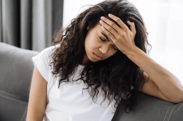 Fáradt fiatal nő ül a kanapén, fáj a feje. A boldogtalan afro-amerikai nő egészségtelen, stresszes, feldúlt a betegség miatt vagy problémái vannak. — Stock Fotó
