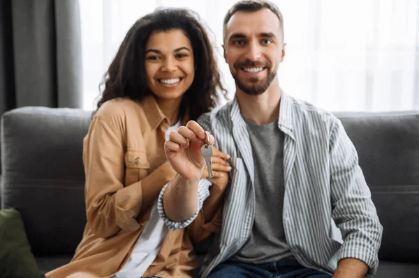 Pareja alegre en un nuevo apartamento o casa, mirando a la cámara y sonriendo. Mujer afroamericana feliz y hombre caucásico sentado en el sofá, sostener una llave en los brazos de su nuevo hogar, día de mudanza —  Fotos de Stock