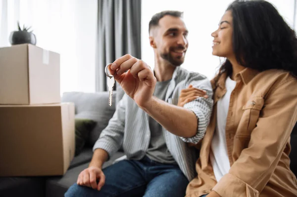 搬到新家去在新公寓或新房子里的一对快乐的夫妇，彼此相视，面带微笑。快乐的非洲裔美国女人和焦虑不安的高加索男人坐在沙发上，手握一把钥匙 — 图库照片