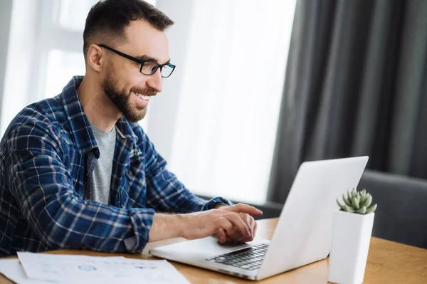 Leende frilansande kille jobbar hemifrån, använder laptop, chattar med kollegor online. Snygg kaukasisk upptagen man utvecklar nya projekt, söker idéer eller planer för start — Stockfoto