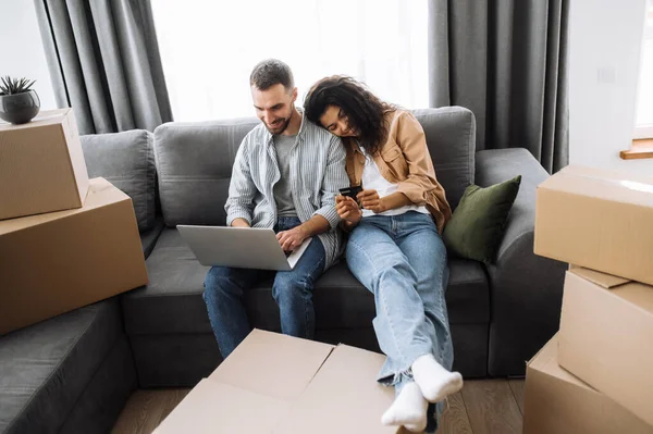 这对快乐的夫妇坐在沙发上，围着纸板箱，搬进新公寓。快乐的高加索男人，带着微笑的美丽的非洲女人，用笔记本电脑，在网上购物，设计了一栋房子 — 图库照片