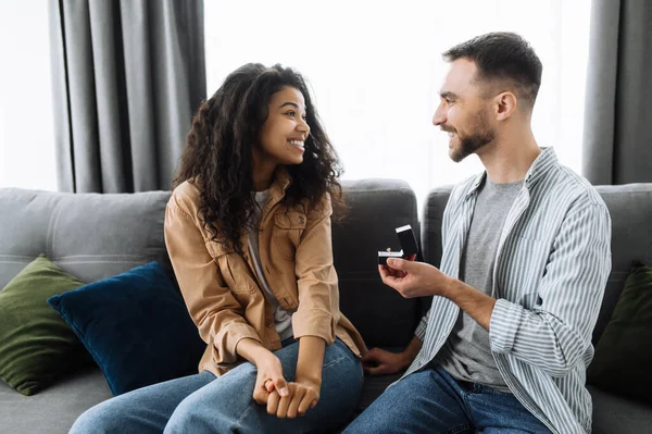 ¿Quieres casarte conmigo? Pareja multirracial enamorada en casa en el sofá en la sala de estar. Caucásico hombre da un anillo de compromiso a su amada afroamericana mujer, ella con gusto lo acepta — Foto de Stock