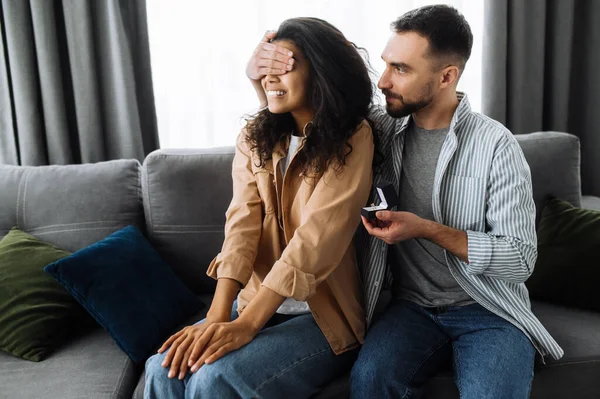 Pareja multiracial enamorada en casa en el sofá del salón. El chico cierra los ojos de su amada novia con su mano, preparándose para darle un anillo y hacerle una oferta de matrimonio — Foto de Stock