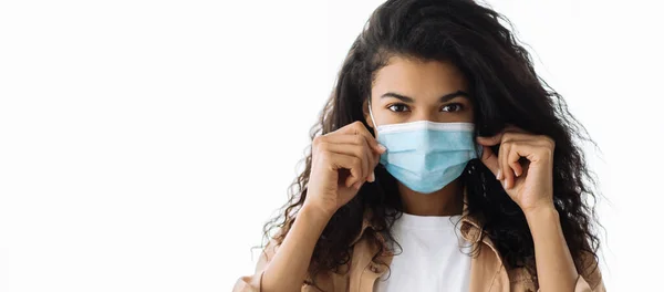 African american brunette woman standing indoors and dress up a protective medical mask on her face and looks at the camera — Stock Photo, Image