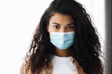 Önleme önlemleri. Afro-amerikan güzel esmer kadın koruyucu bir maskeyle evde dikiliyor ve kameraya bakıyor.