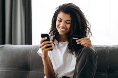 Kadının elinde bir kredi kartı ve akıllı telefon. Neşeli Afrikalı Amerikalı genç kadın telefon ekranına bakıyor, internetten alışveriş yapıyor, online ödeme yapıyor.