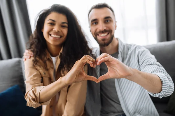 Un gesto cardíaco en primer plano. Desenfocados, una pareja enamorada se sienta en el sofá y muestra un gesto de corazón con sus manos. Concepto de amor y relación — Foto de Stock