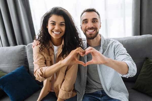 Una pareja multirracial enamorada se sienta en el sofá de su casa y muestra un gesto de corazón con las manos. Concepto de amor y relación — Foto de Stock