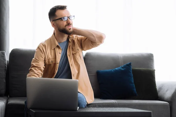 Kaukasiska manliga frilansare killen sitter på soffan hemma, med laptop. Trött programmerare utveckla ett projekt, har ont i ryggen, behöver vila eller paus — Stockfoto