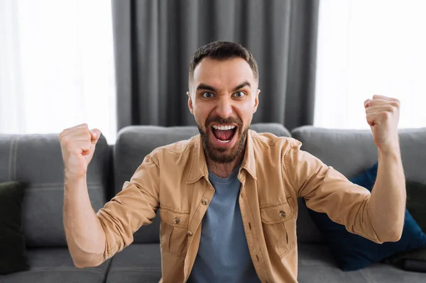 Emotional screaming man watching tv, support and cheering for favorite team. Excited cheerful male sits on a sofa at home, celebrate results of match, or got a great news or deal