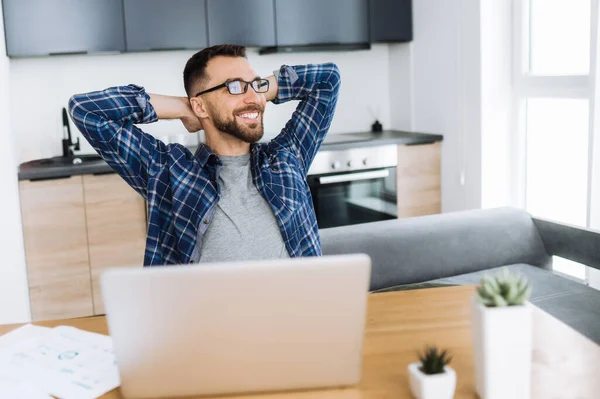 Positiv frilansande kille som kopplar av på arbetsplatsen och ler. Självsäker kaukasisk manlig entreprenör sitter på hemmakontoret, tar paus från distansarbete och drömmer om semester — Stockfoto