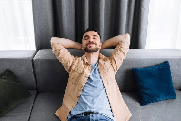 Ελκυστικός άνθρωπος χαλαρώνει στον καναπέ στο σαλόνι στο σπίτι και χαμογελώντας. Καυκάσιος κομψός τύπος κάνει ένα διάλειμμα από τη δουλειά στο διαδίκτυο, ή έχει Σαββατοκύριακο, ονειρεύεται διακοπές — Φωτογραφία Αρχείου