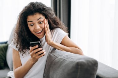 Heyecanlı güzel kadın akıllı telefon kullanıyor, ekrana şaşırmış görünüyor. Kanepede oturan Afrikalı Amerikalı genç bir kadın iyi bir haber ya da mesaj getirdi.