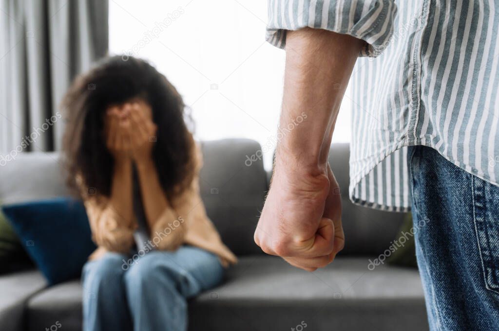 Aggression by a man to his woman, a man clenched his fist to hit his wife or girlfriend, the girl in defocus sitting on the couch covering herself with her hands, concept of domestic violence to