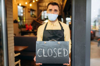 Üzgün garson, kahveci ya da küçük işletme sahibi. Tıbbi koruyucu maskeli mutsuz bir adam ve siyah iş önlüğü bir kafe, restoran ya da barın girişinde duruyor ve elinde kapalı işareti var.