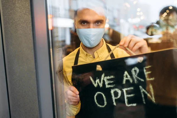 Welcome. Satisfied caucasian male waiter or barista showing signboard OPEN while standing at restaurant or cafe entrance wearing medical mask on his face and black apron and invites clients